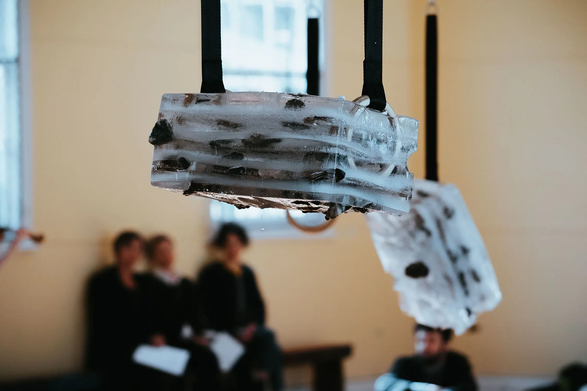 In a gallery space, frozen iceblock sculptures are suspended by black straps and metal hoops frozen into the ice. People sit on a bench against the wall in the background.