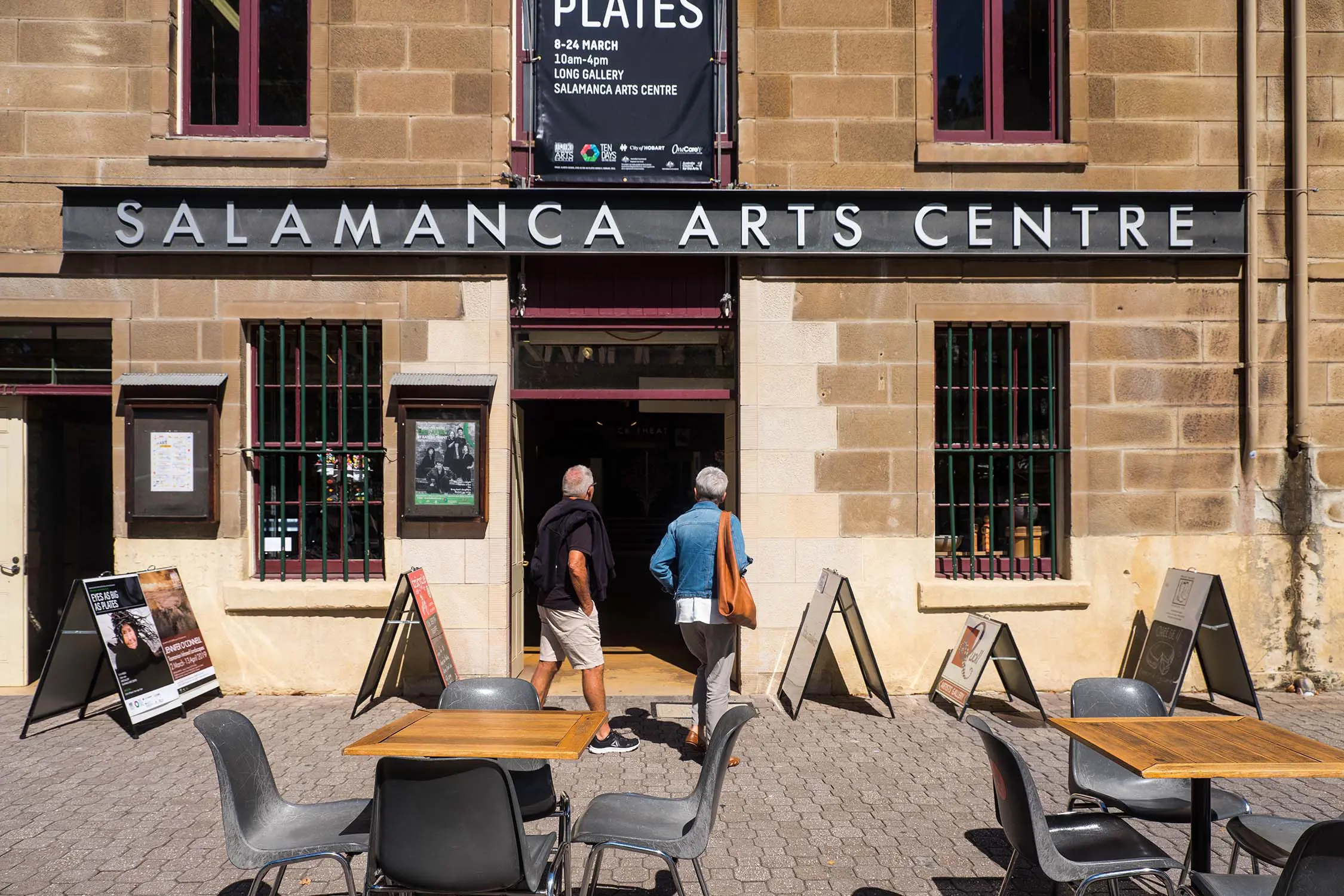 Two people step into an entrance of a historic sandstone building, with the sign above the door reading "Salamanca Arts Centre". Tables, chairs and sandwich boards line the footpath outside.