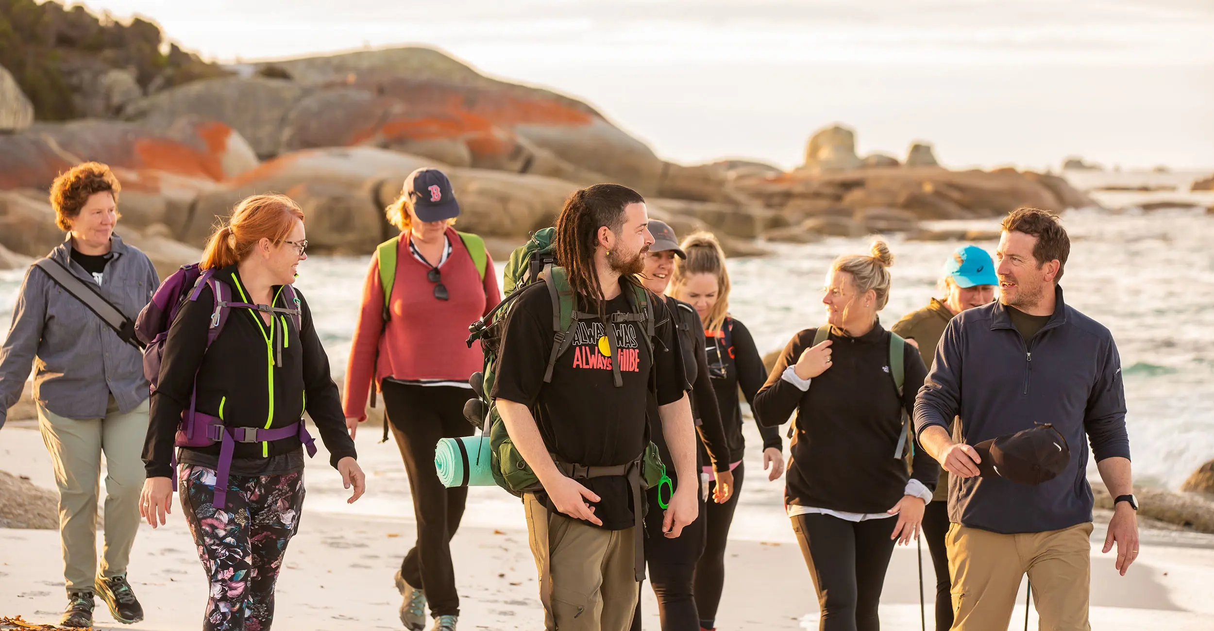 A group of people are led by a guide across white sands in front of orange rocks on a beach.