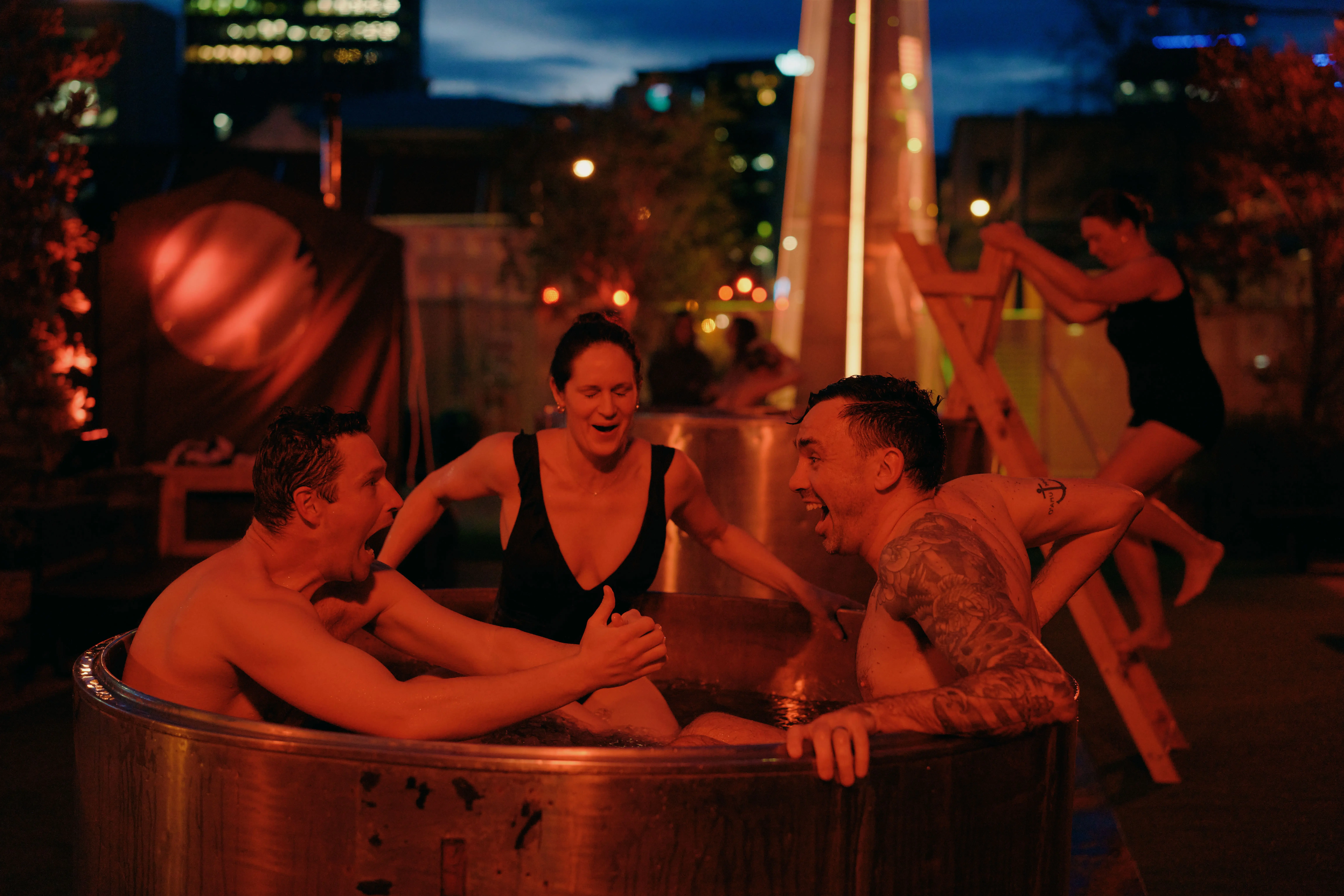 Two men and a woman in swimsuits yell and smile while sitting in a round metal bath outdoors, the scene is bathed in red light. lower themselves into a