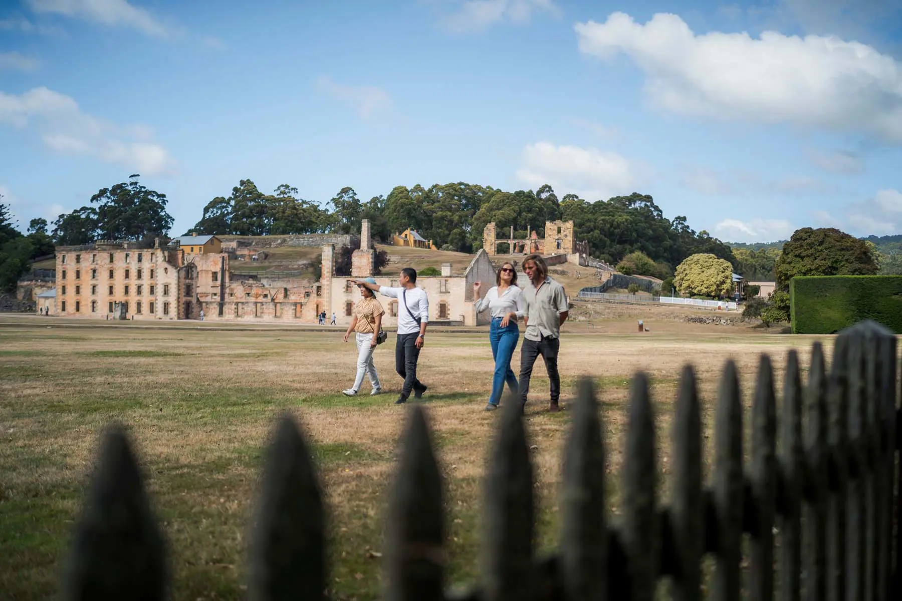 Four people walk across grass in front of a black wrought iron fence. Behind them, historic sandstone buildings of a convict site sit against a bush-covered hill.