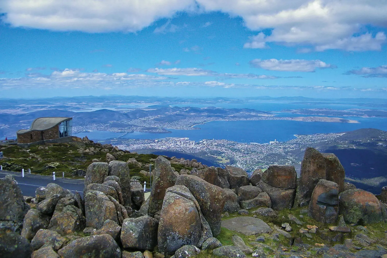 A view over the lookout area atop a mountain. Large boulders are dotted nearby a lookout shelter, and the view is over Hobart and the Derwent River.
