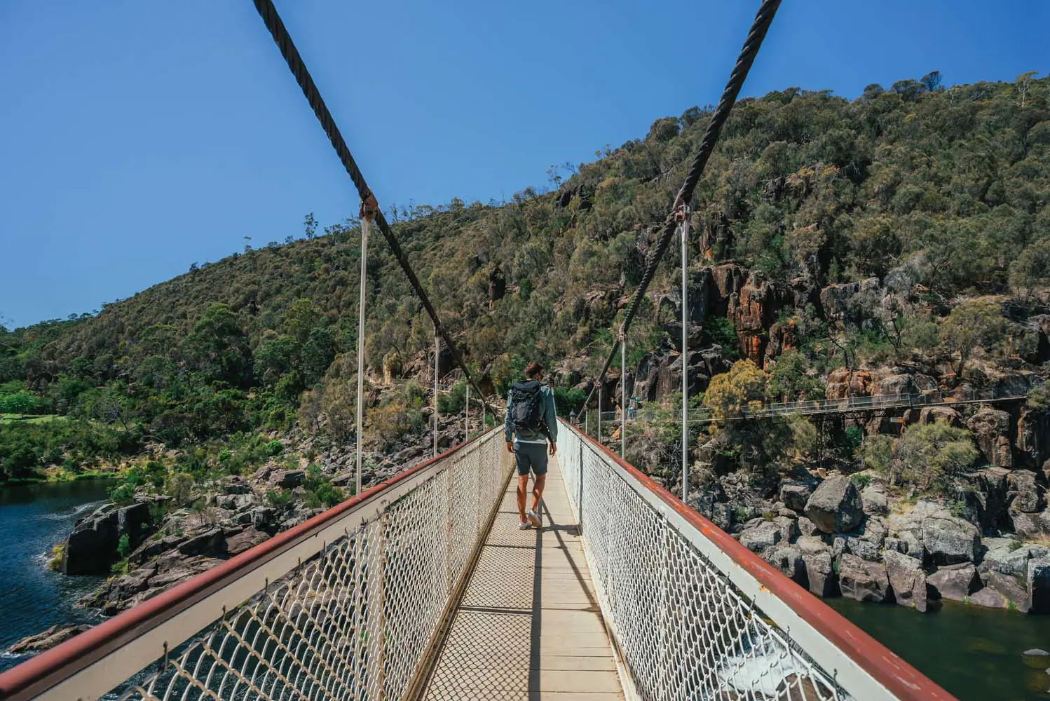 A person walking away down a long suspension bridge, heading into the bushy cliffs on the other side.