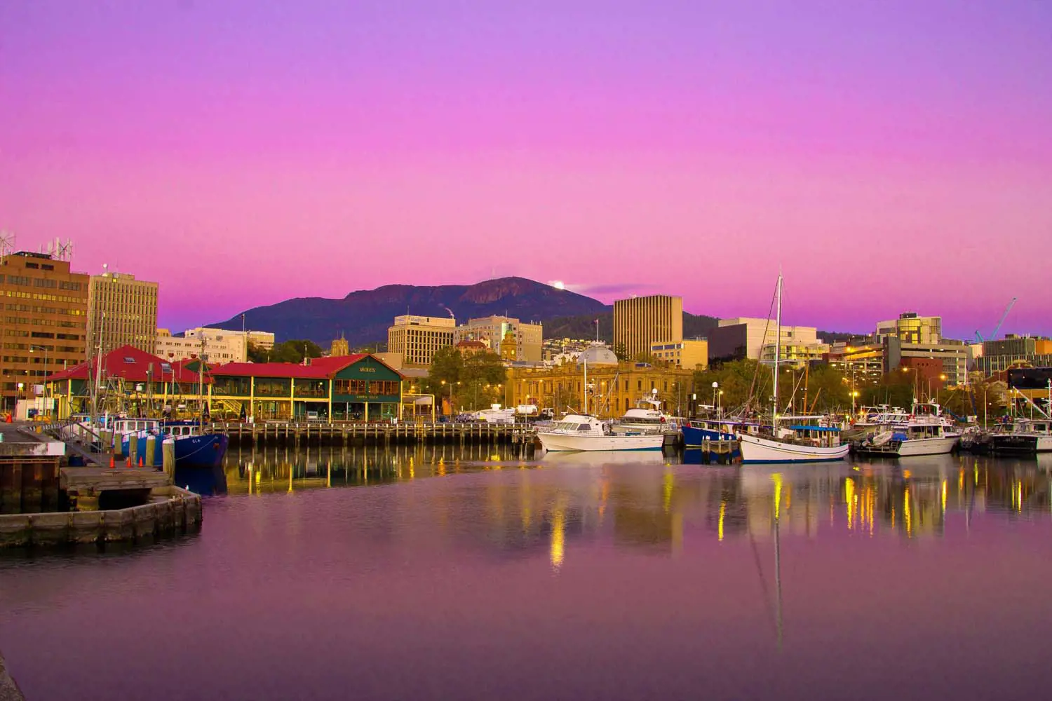 A pink and purple sunset shines down on a waterfront marina area, with moored boats and heritage sandstone buildings visible.