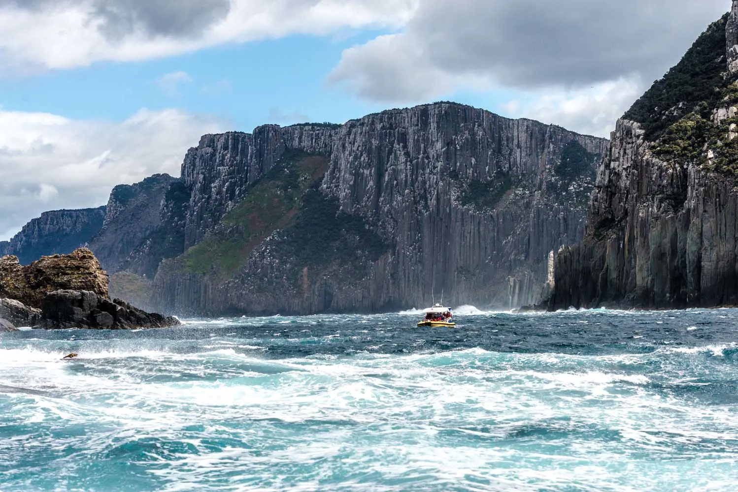 A small boat cruises through choppy water at the base of sharply rising, imposingly dark cliffs.