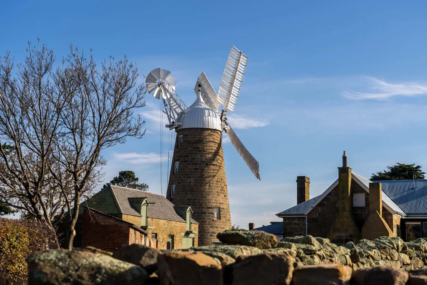A picturesque image of a heritage windmill and brick buildings, standing tall against a bright blue sky with a few wisps of cloud.