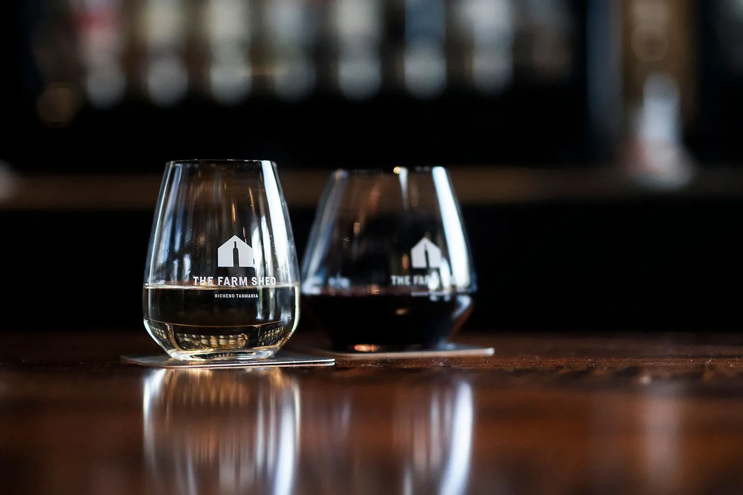 Two stemless wineglasses - one with red, one with white - sit on coasters on a rich dark wood table.