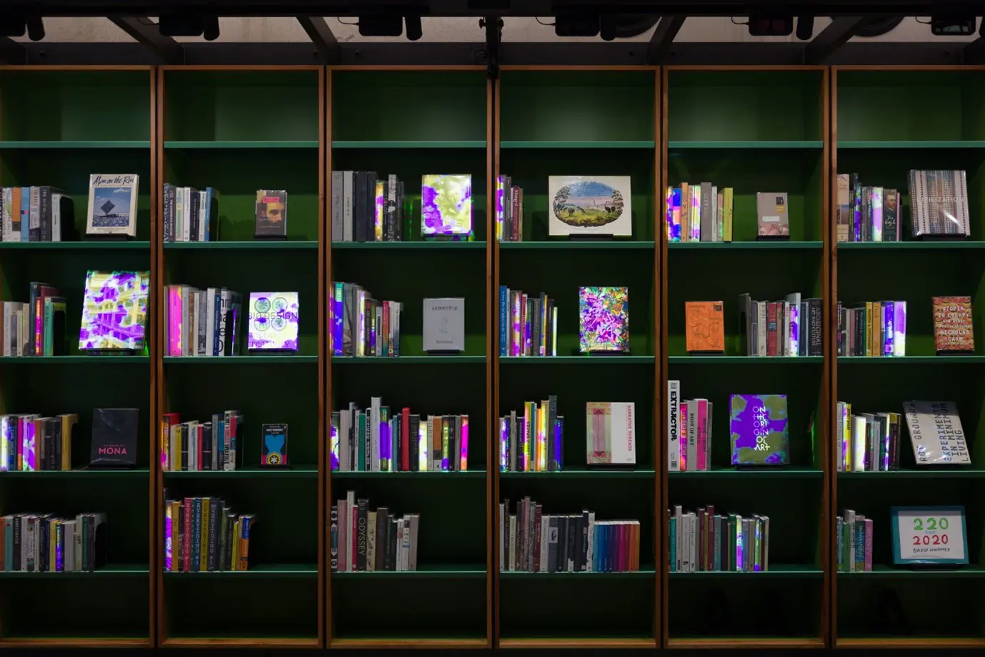 Neat, ceiling-height dark green shelves fully line a wall. Each shelf has an array of books, some on stands facing to the front.