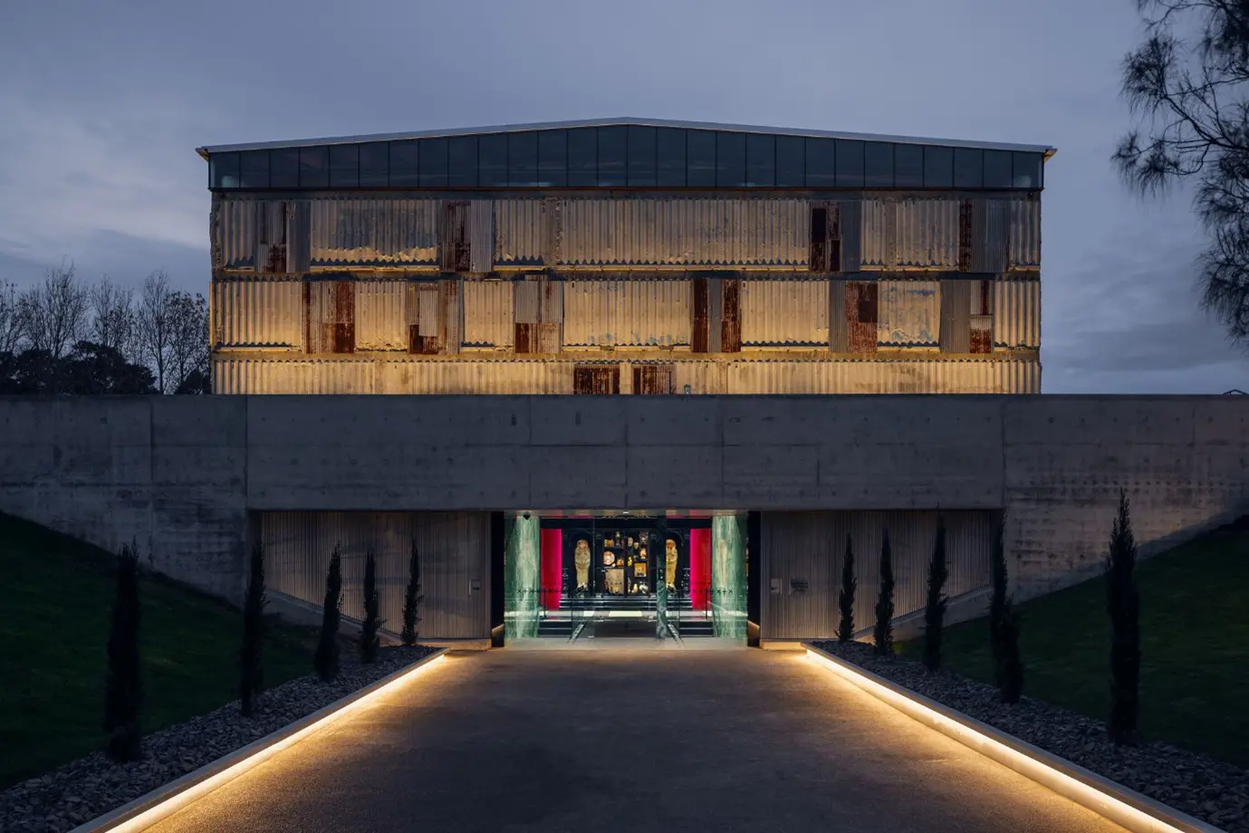 A tall, imposing concrete building at dusk. Illuminated pathways lead towards a colourfully lit entrance, while a huge concrete building looms overhead.
