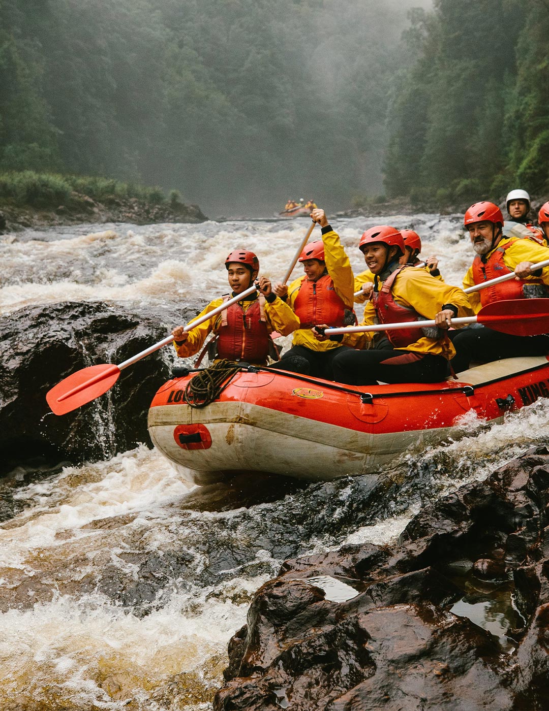 A group of rafters in brightly-coloured safety gear and helmets paddle their raft over fast moving rapids between rocks. 