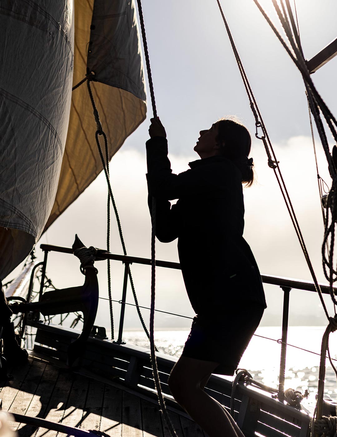 A woman stands in silhouette, pulling at a rope securing sails on a large sailing boat.